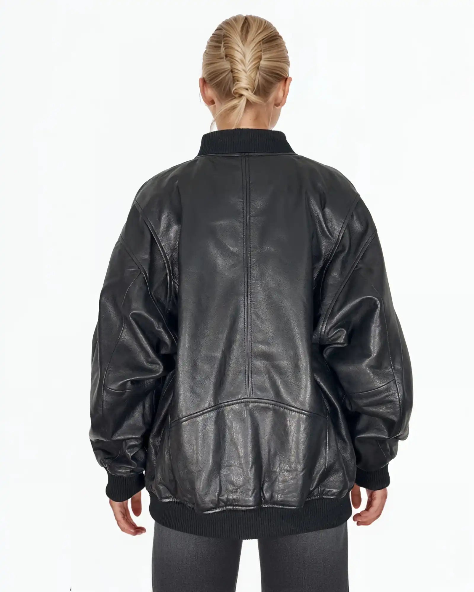 Eine Person mit blondem Haar in einem tiefen Zopf ist von hinten zu sehen. Sie trägt die trueprodigy Selina Oversize-Bomberjacke aus Leder mit Rippbündchen & schwarzem Zipper, gepaart mit einer dunklen Hose vor einem schlichten weißen Hintergrund.