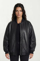 Eine Frau mit langen dunklen Haaren steht mit dem Gesicht nach vorne und trägt eine trueprodigy Oversize Bomber Lederjacke Damen "Selina" über einem schwarzen Oberteil und einer schwarzen Hose vor einem schlichten hellen Hintergrund.