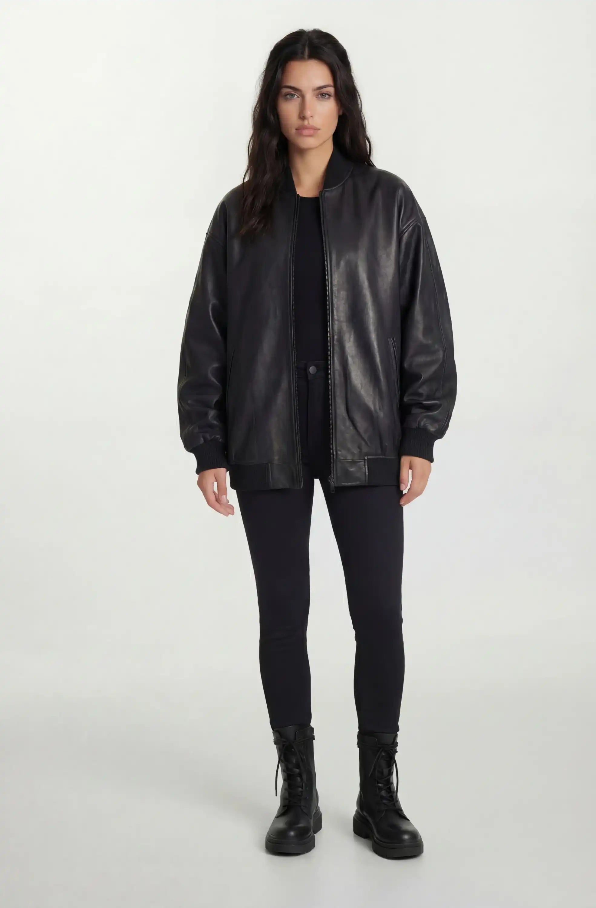 Eine Frau steht vor einem schlichten, hellen Hintergrund und trägt die trueprodigy Oversize-Bomberjacke aus Leder mit Rippbündchen & schwarzem Reißverschluss, gepaart mit einem schwarzen Hemd, Skinny Jeans und Schnürstiefeln, die Arme an den Seiten.