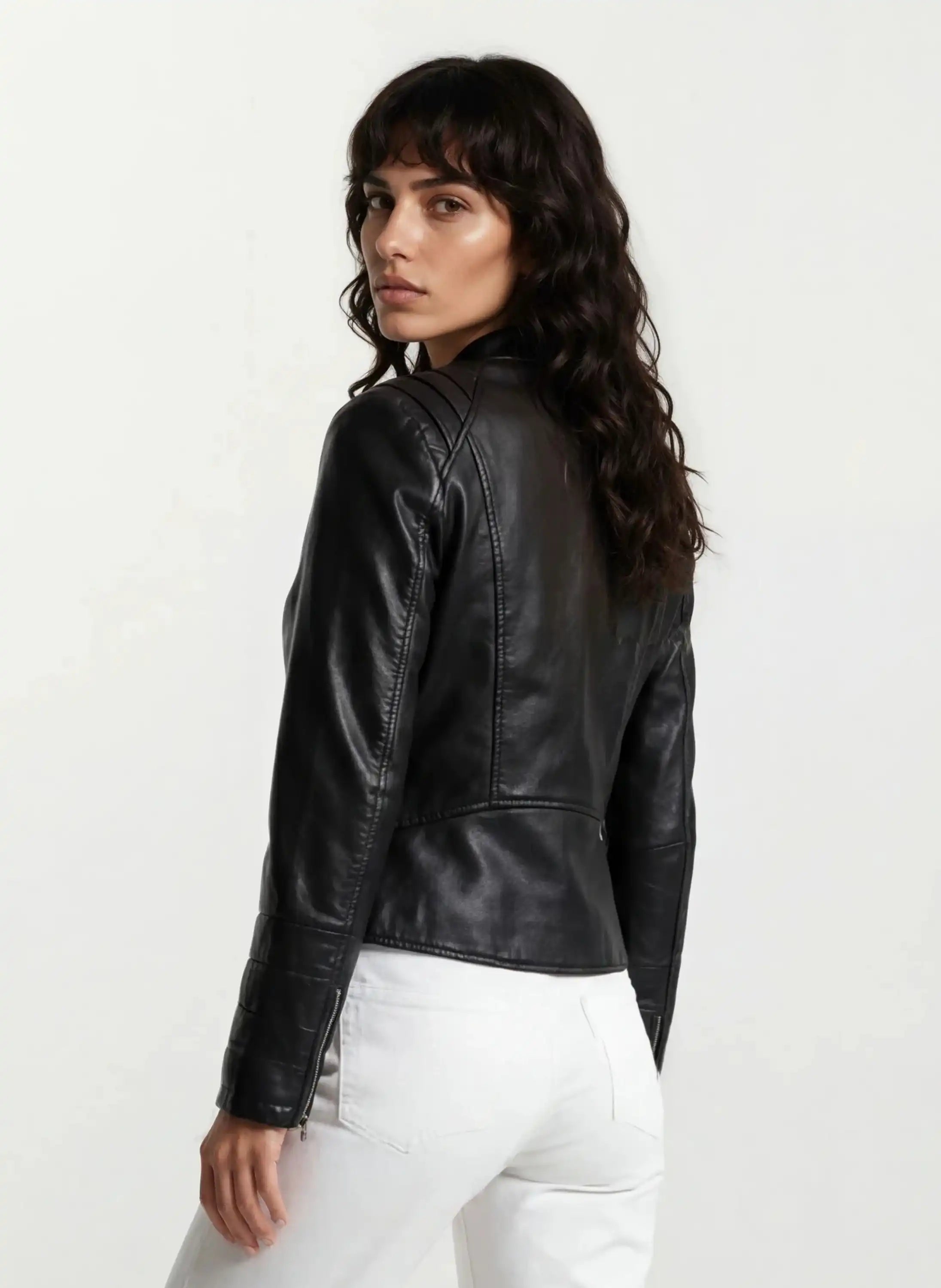 Eine Frau mit langen, gewellten braunen Haaren steht seitlich in weißen Hosen und der trueprodigy Taillierte Lederjacke mit Stehkragen, Steppnähten & silbernen Reißverschlüssen vor einem hellen Hintergrund.