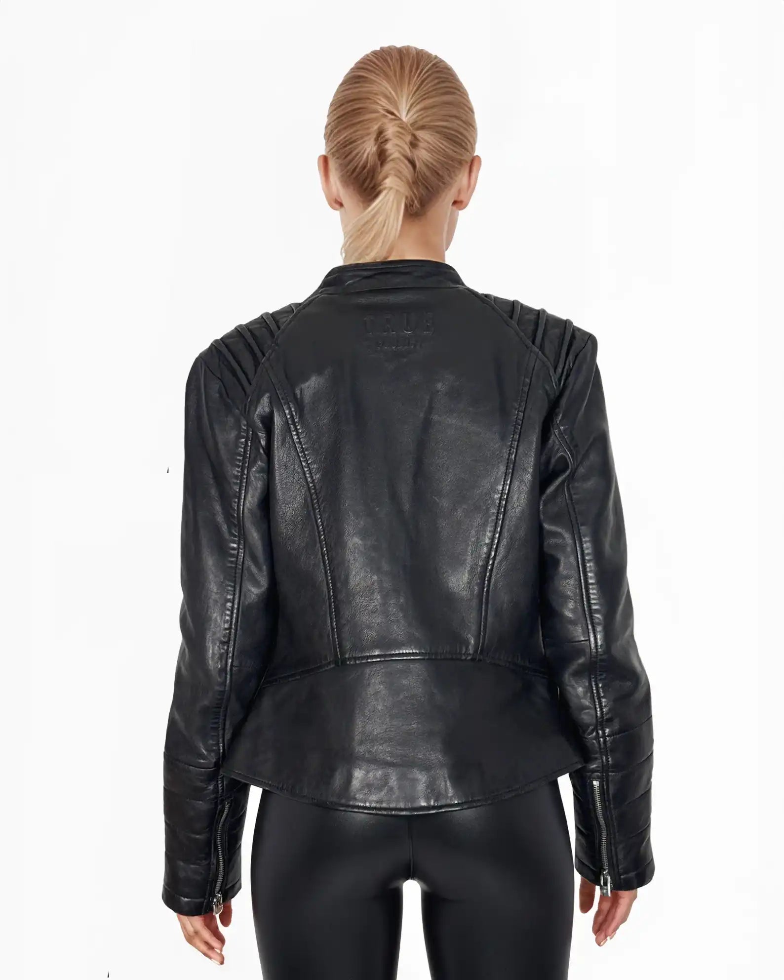 Eine Person mit blonden, geflochtenen Haaren trägt von hinten sichtbar die schwarze Raegan Taillierte Lederjacke mit Stehkragen, Steppnähten & silbernen Reißverschlüssen von trueprodigy sowie eine schwarze Hose vor weißem Hintergrund.