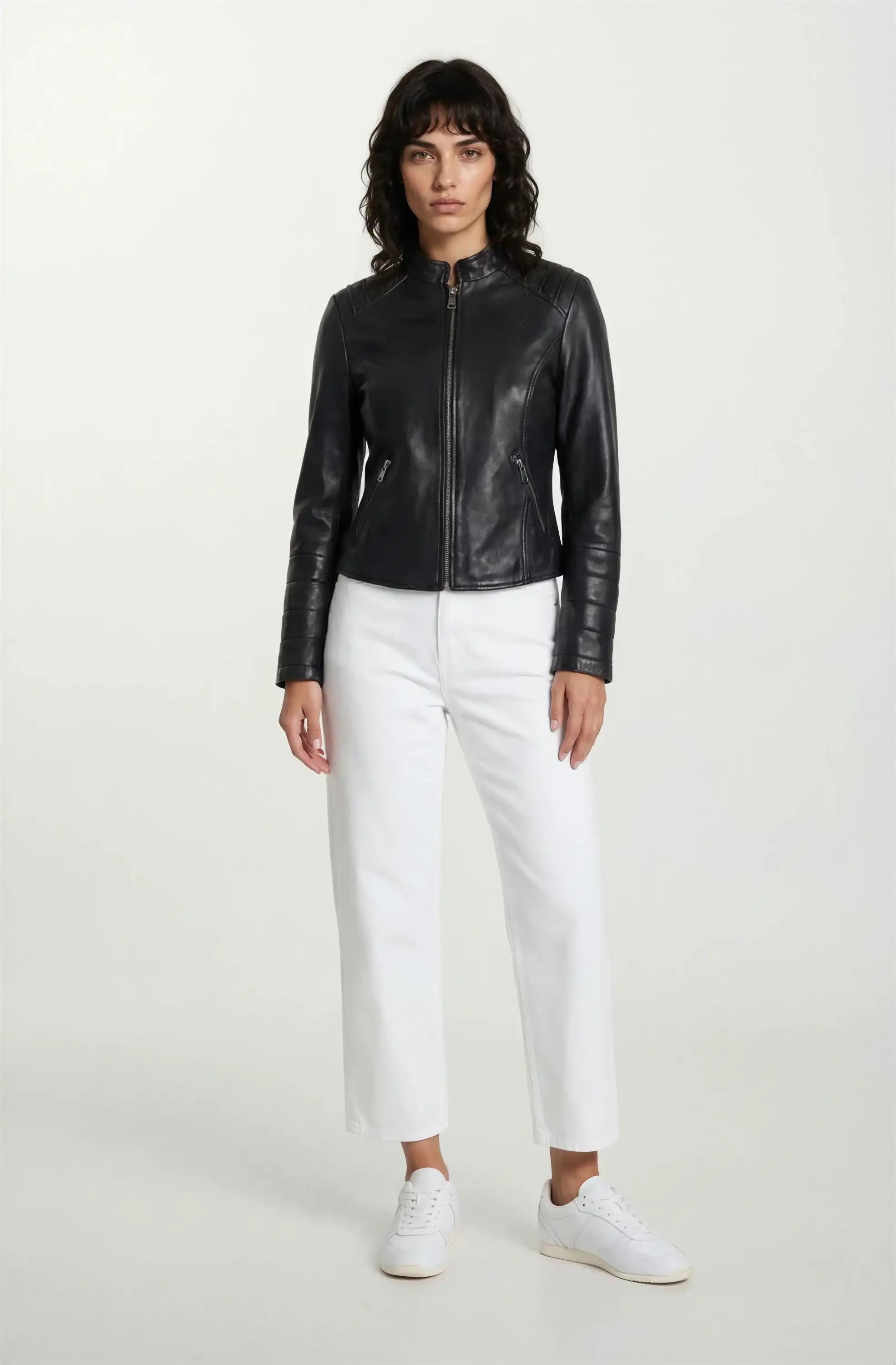 Eine Person blickt nach vorne und trägt eine schwarze trueprodigy Taillierte Lederjacke mit Stehkragen, Steppnähten & silbernen Reißverschlüssen, eine weiße Hose und weiße Turnschuhe vor einem schlichten weißen Hintergrund.