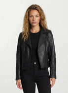 Eine Frau mit langen, welligen blonden Haaren trägt die taillierte Lederjacke von trueprodigy mit schwarzem Reißverschluss und Ärmelzipper über schwarzem Shirt und Hose. Sie steht vor hellem Hintergrund und blickt neutral in die Kamera.