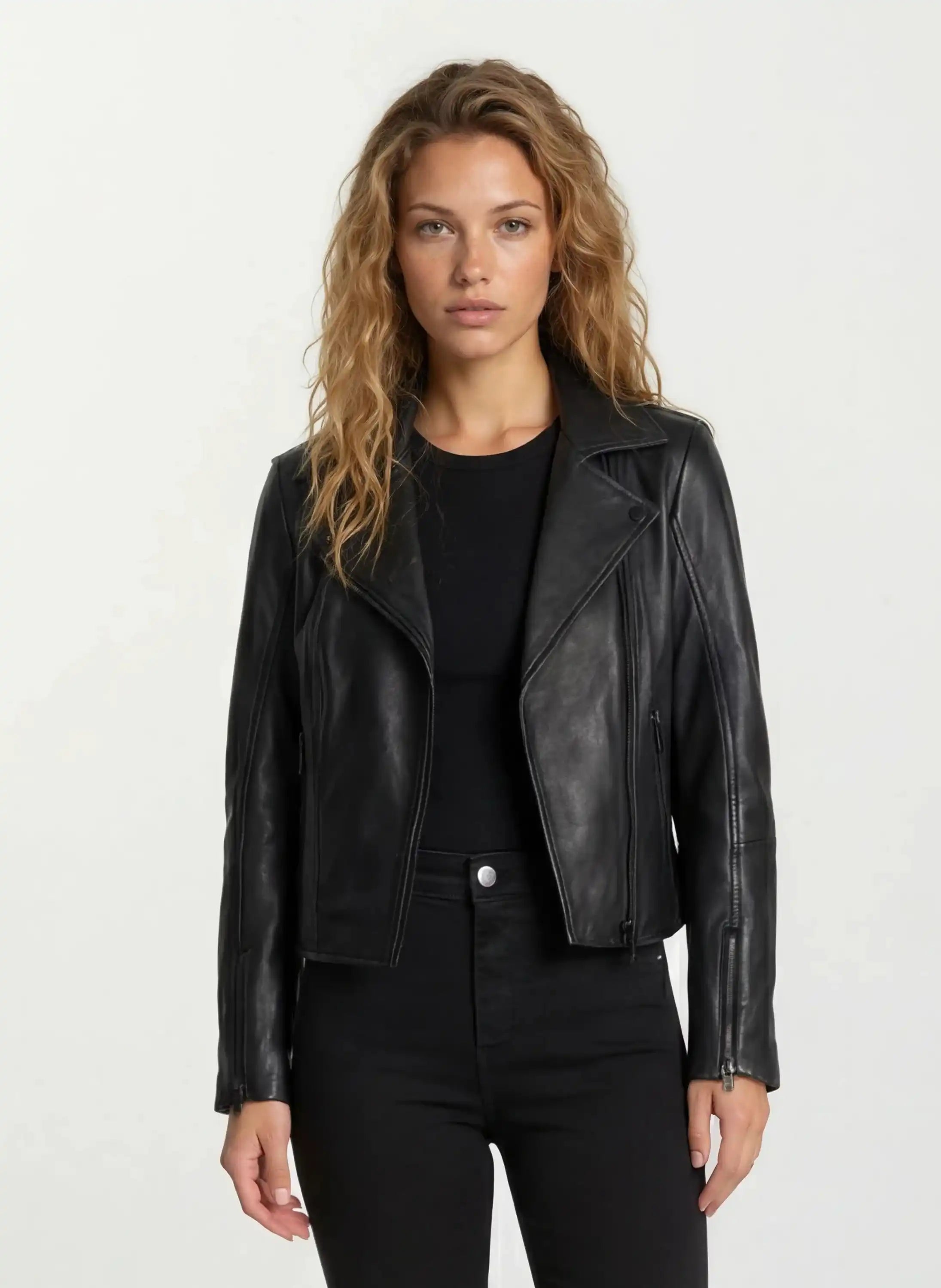Eine Frau mit langen, welligen blonden Haaren trägt die taillierte Lederjacke von trueprodigy mit schwarzem Reißverschluss und Ärmelzipper über schwarzem Shirt und Hose. Sie steht vor hellem Hintergrund und blickt neutral in die Kamera.