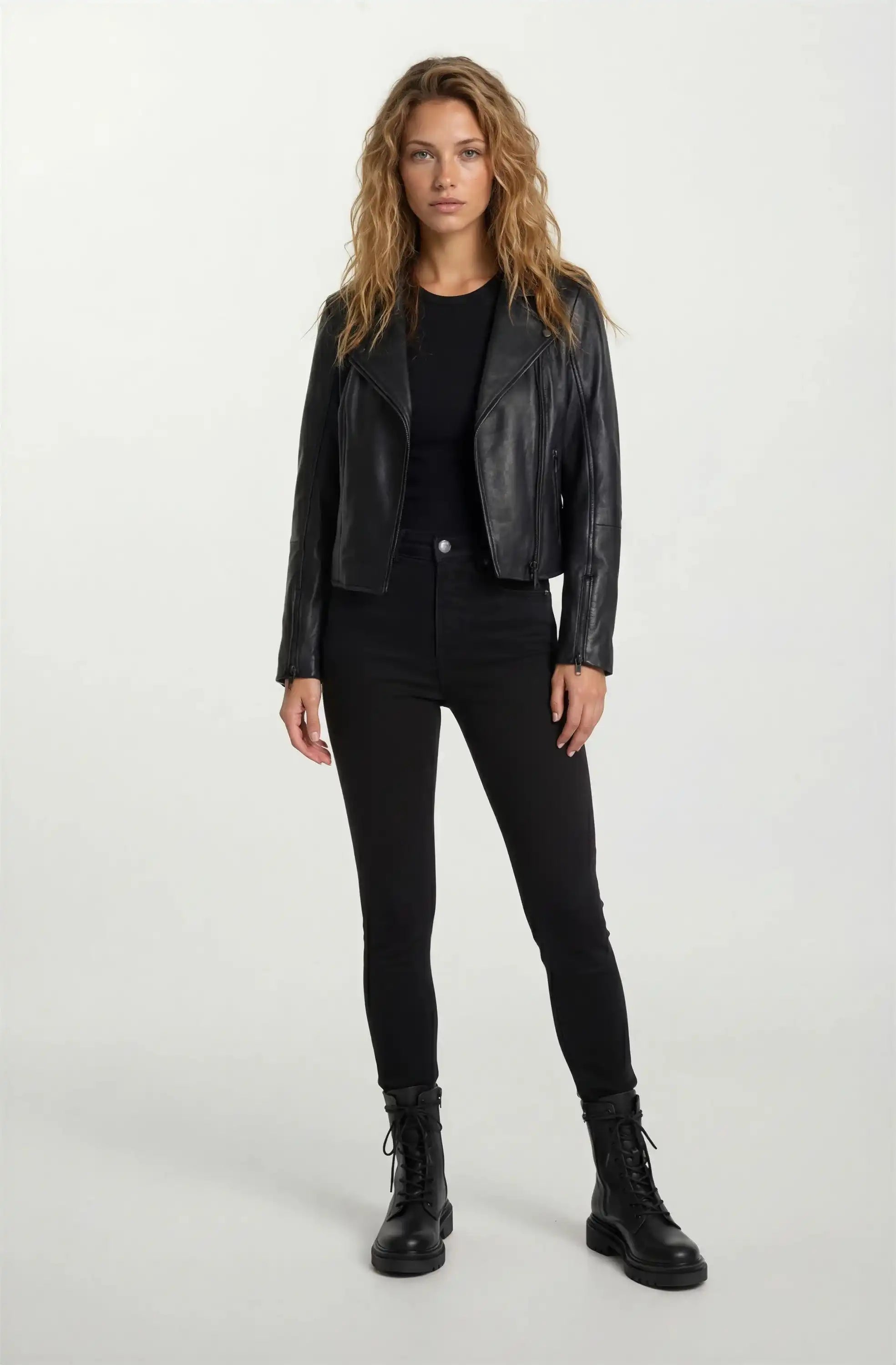 Eine Frau mit langem, gewelltem Haar steht mit dem Gesicht nach vorne und trägt eine taillierte trueprodigy Lederjacke mit schwarzem Reißverschluss & Ärmelzipper, schwarzem Shirt, Skinny Jeans und Schnürboots vor hellem Hintergrund.