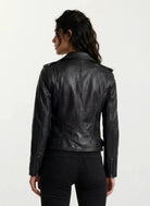 Eine Person mit gewelltem dunklem Haar ist von hinten zu sehen. Sie trägt eine taillierte trueprodigy Feminine Bikerjacke mit Gürtel & Schulterriegeln und schwarze Jeans vor einem schlichten hellen Hintergrund.
