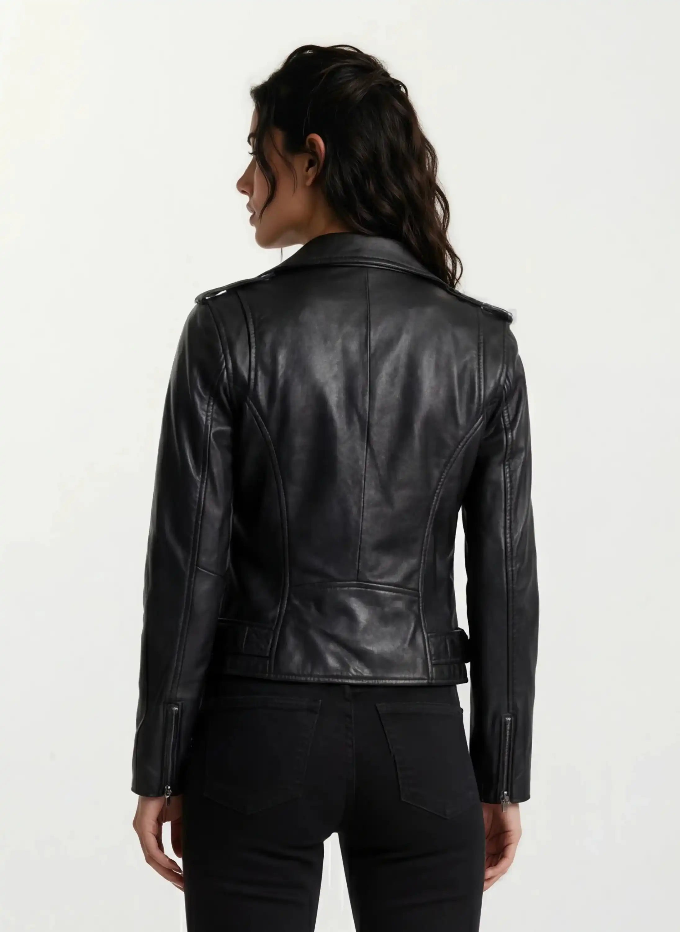 Eine Person mit gewelltem dunklem Haar ist von hinten zu sehen. Sie trägt eine taillierte trueprodigy Feminine Bikerjacke mit Gürtel & Schulterriegeln und schwarze Jeans vor einem schlichten hellen Hintergrund.