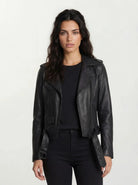 Eine Frau mit langen dunklen Haaren steht vor der Kamera und trägt eine trueprodigy Feminine Bikerjacke mit Gürtel & Schulterriegeln über einem schwarzen Hemd und einer Hose vor einem schlichten hellen Hintergrund.