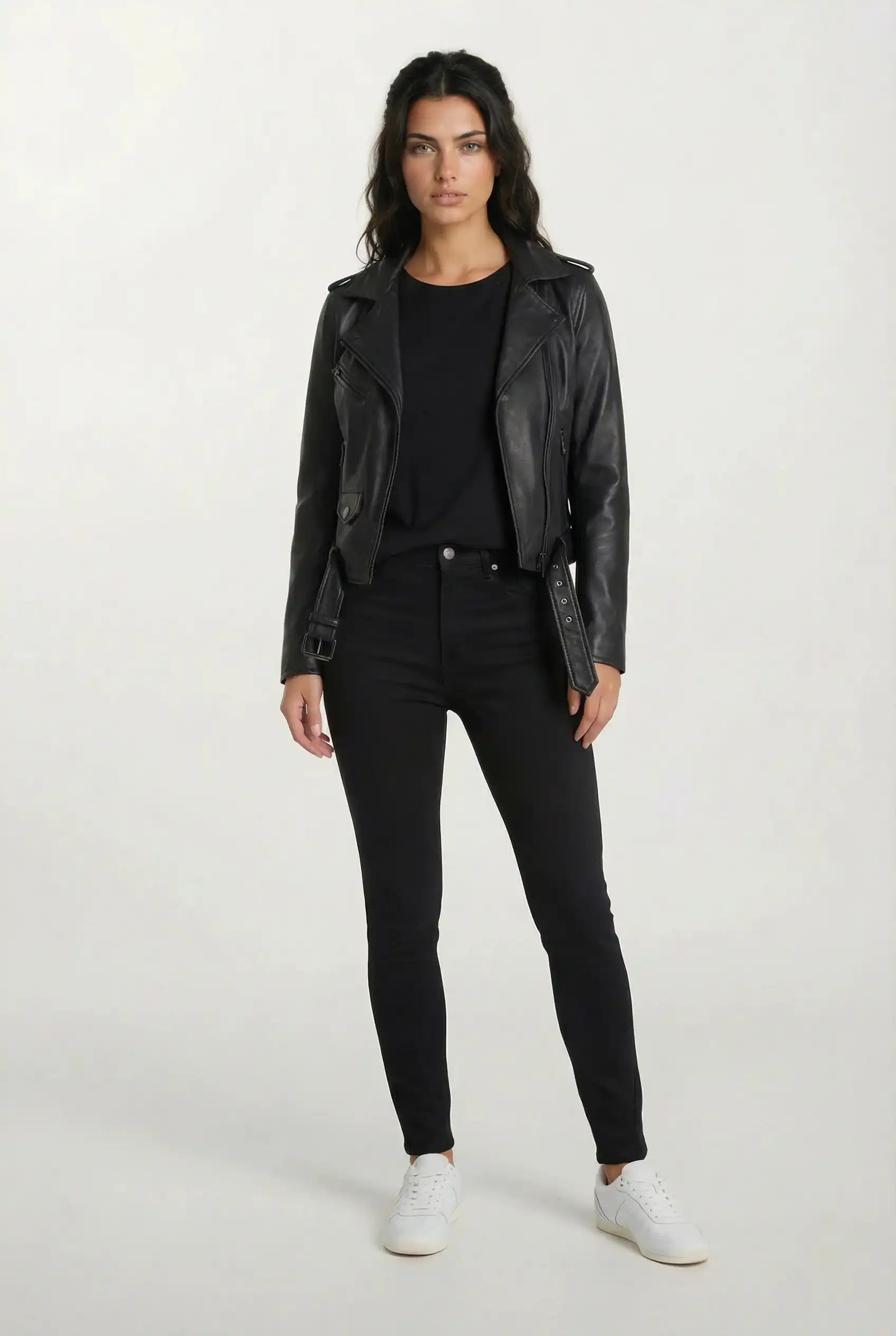 Vor einem hellen Hintergrund trägt eine Frau die trueprodigy Feminine Bikerjacke mit Gürtel & Schulterriegeln, ein schwarzes Top, schwarze Jeans und weiße Sneaker. Sie hat lange dunkle Haare und einen neutralen Gesichtsausdruck.