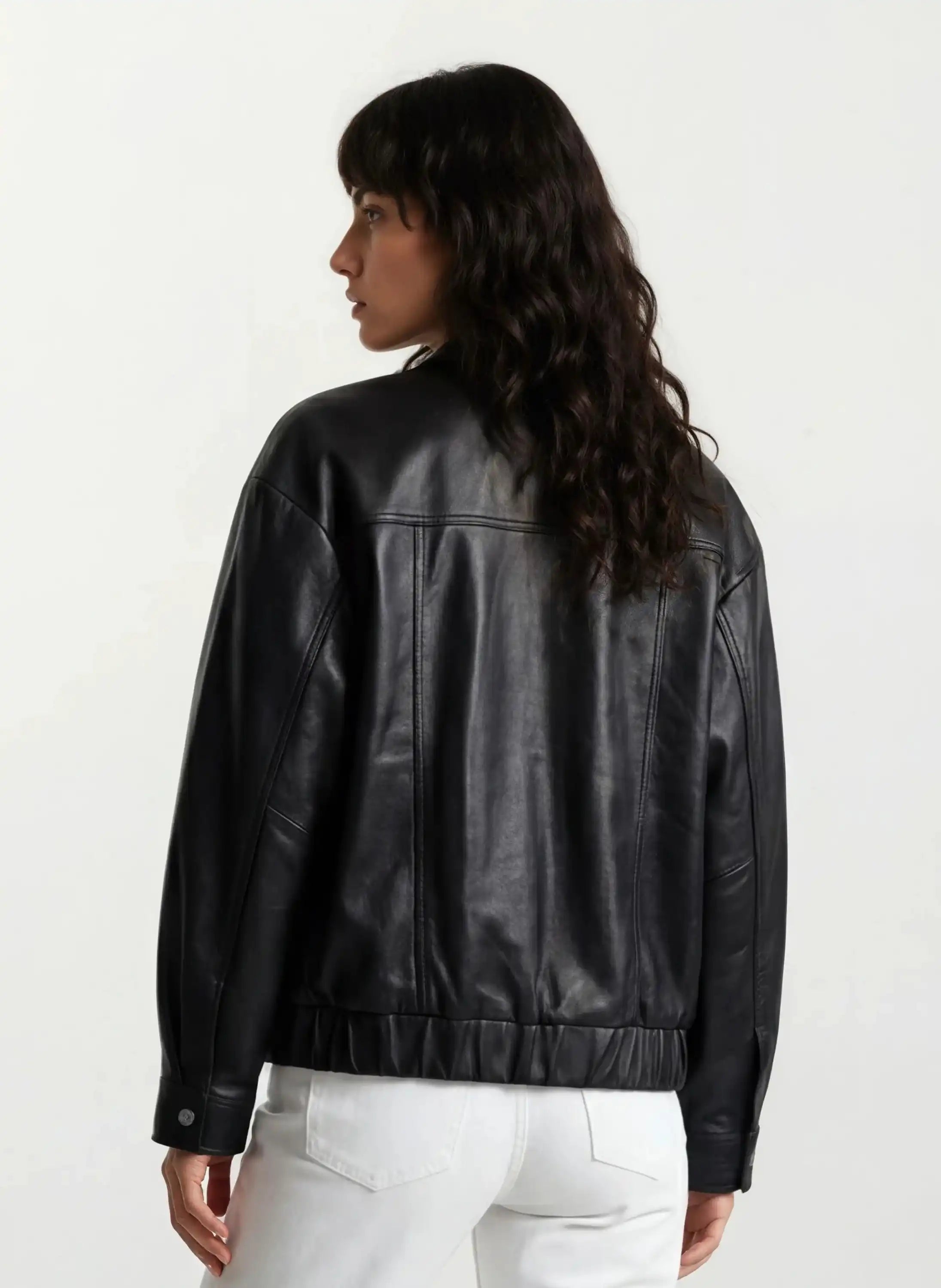 Eine Person mit langen, gewellten dunklen Haaren wird von hinten gezeigt. Sie trägt eine trueprodigy Oversize-Lederjacke mit Gummizugsaum & Lederdetails und eine weiße Hose vor einem schlichten weißen Hintergrund.