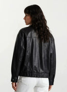 Eine Person mit langen, gewellten dunklen Haaren wird von hinten gezeigt. Sie trägt eine trueprodigy Oversize-Lederjacke mit Gummizugsaum & Lederdetails und eine weiße Hose vor einem schlichten weißen Hintergrund.