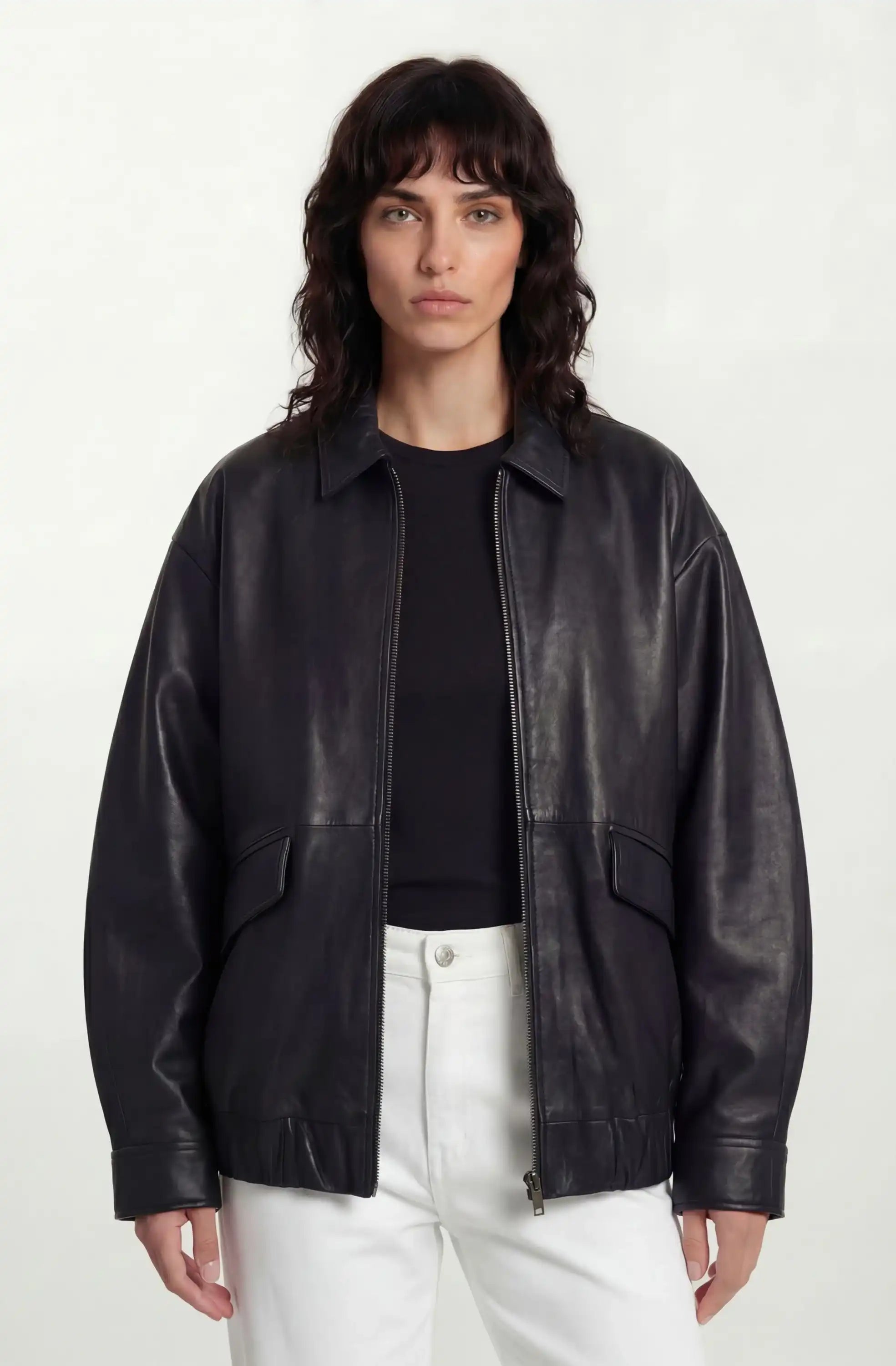 Eine Frau mit gewelltem, schulterlangem dunklem Haar trägt eine trueprodigy Oversize-Lederjacke mit Gummizugsaum & Lederdetails über einem schwarzen Oberteil und einer weißen Hose, posiert vor einem schlichten hellen Hintergrund.