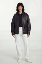 Eine Frau mit mittellangem dunklem Haar trägt die trueprodigy Oversize-Lederjacke mit Gummizugsaum & Lederdetails, ein schwarzes Oberteil, eine weiße Hose und weiße Schuhe. Sie steht vor einem schlichten weißen Hintergrund und blickt mit neutralem Blick nach vorne.