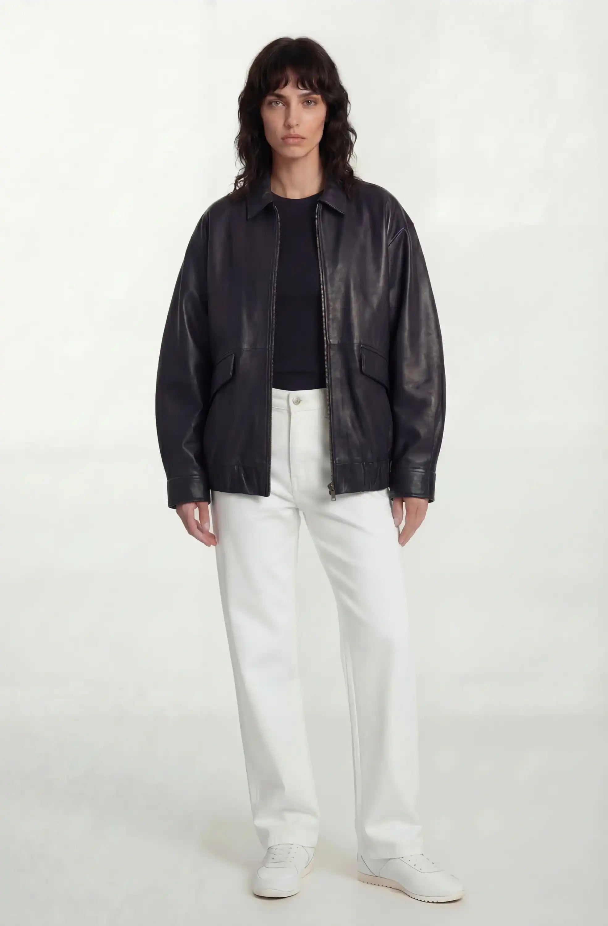 Eine Frau mit mittellangem dunklem Haar trägt die trueprodigy Oversize-Lederjacke mit Gummizugsaum & Lederdetails, ein schwarzes Oberteil, eine weiße Hose und weiße Schuhe. Sie steht vor einem schlichten weißen Hintergrund und blickt mit neutralem Blick nach vorne.