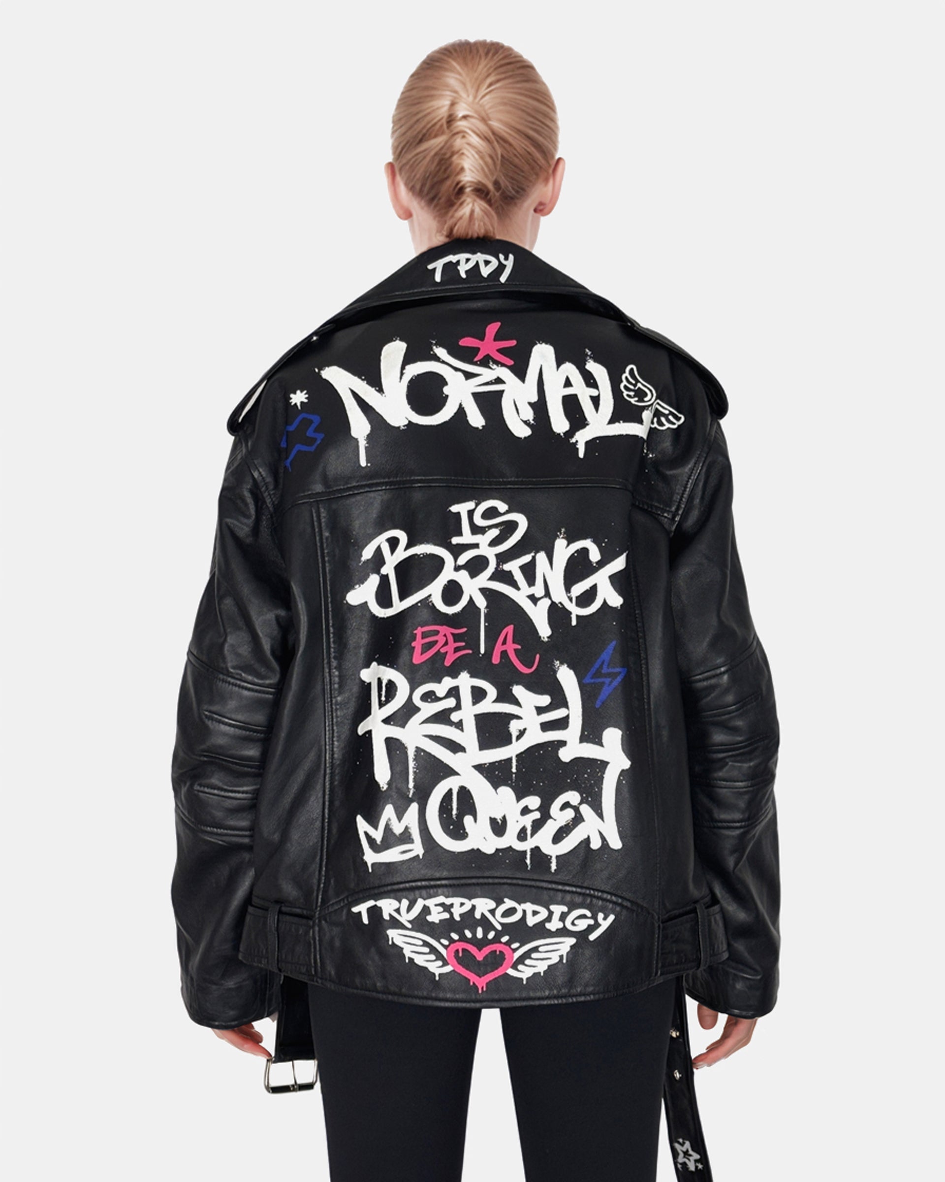 Eine blonde Person mit Pferdeschwanz trägt die „Anya“ schwarze Graffiti-Lederjacke von trueprodigy mit weißem Rückenaufdruck „Normal is langweilig, be a rebel queen“, Symbole und einem punkigen Touch.