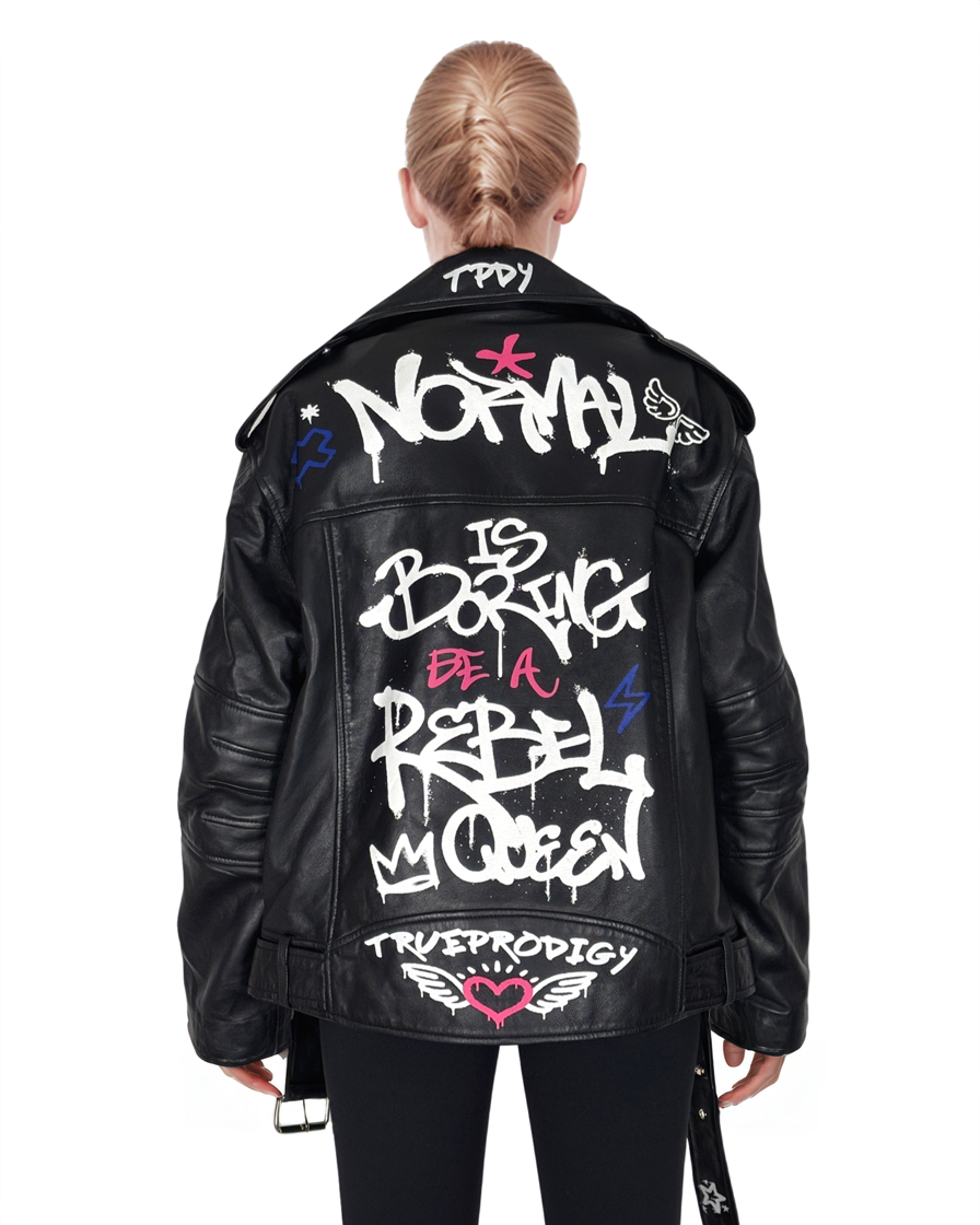Eine blonde Person mit Zopf trägt die Schafslederjacke „Anya“ von trueprodigy mit weißem Graffiti-Text – „Normal ist langweilig, sei eine Rebellenkönigin, trueprodigy“ – und verschiedenen grafischen Symbolen.