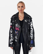 Eine Person trägt die schwarze Graffiti-Lederjacke „Anya“ von trueprodigy aus Schafsleder mit bunten Designs, kombiniert mit schwarzen Leggings und schwarzem Top – für einen punkigen Look vor weißem Hintergrund.
