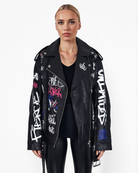 Eine Frau mit zurückgekämmten blonden Haaren präsentiert die Graffiti-Jacke „Anya“ von Trueprodigy aus Schafsleder mit bunten Schriftzügen und Symbolen, kombiniert mit einem schwarzen Oberteil und glänzenden schwarzen Hosen, vor einem weißen Hintergrund.