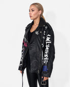 Eine Frau mit zurückgebundenem blonden Haar trägt die Graffiti-Lederjacke „Anya“ von trueprodigy aus Schafsleder mit weißen und farbigen Textdetails über einem schwarzen Oberteil und einer glänzenden schwarzen Hose, die vor einem schlichten weißen Hintergrund einen Punk-Touch verleiht.