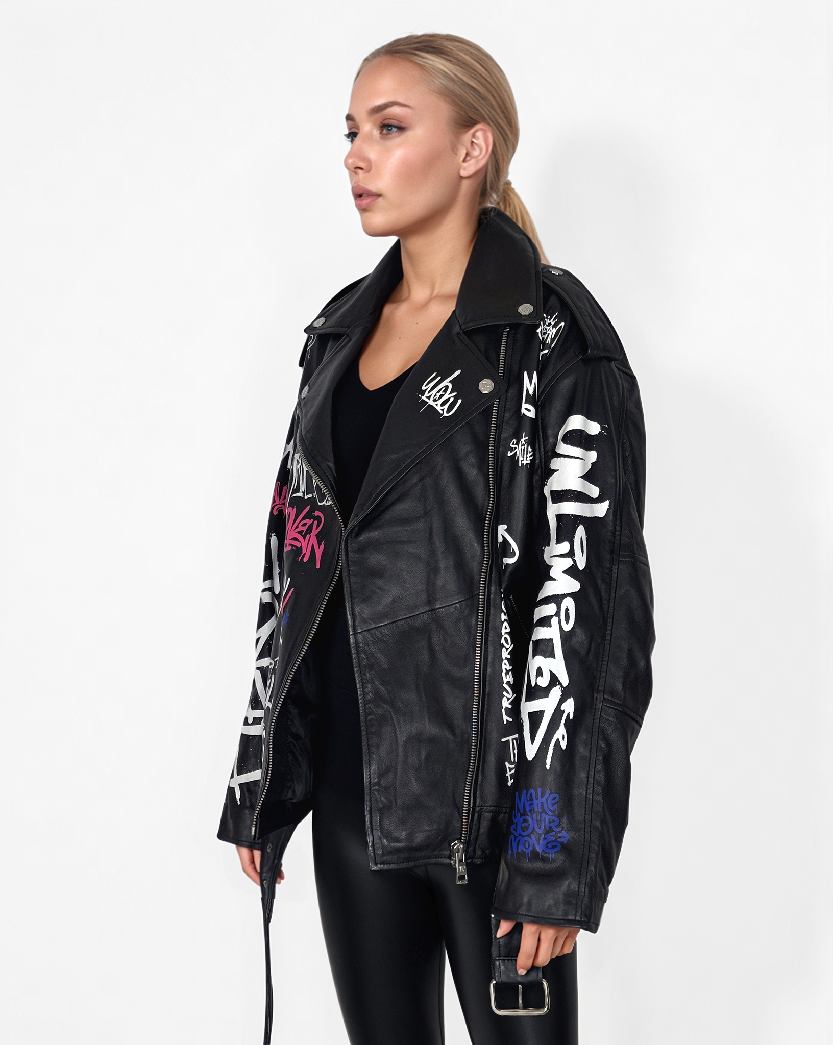 Eine blonde Frau steht vor einem weißen Hintergrund und trägt die schwarze übergroße Graffiti-Lederjacke „Anya“ von trueprodigy mit weißem und buntem Text, einem stylischen Reverskragen und schwarzen Leggings.