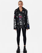 Vor einem schlichten weißen Hintergrund steht eine Frau, die die Schafslederjacke „Anya“ von Trueprodigy mit weißen, blauen und roten Graffiti-Designs trägt, kombiniert mit schwarzen Leggings und Plateaustiefeln für einen Punk-inspirierten Look.