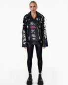 Eine Frau steht vor weißem Hintergrund und trägt die schwarze Graffiti-Lederjacke „Anya“ mit Reverskragen aus Schafsleder von trueprodigy, dazu schwarze Leggings und Plateau-Stiefel. Sie hat blonde, zurückgebundene Haare und einen neutralen Gesichtsausdruck.
