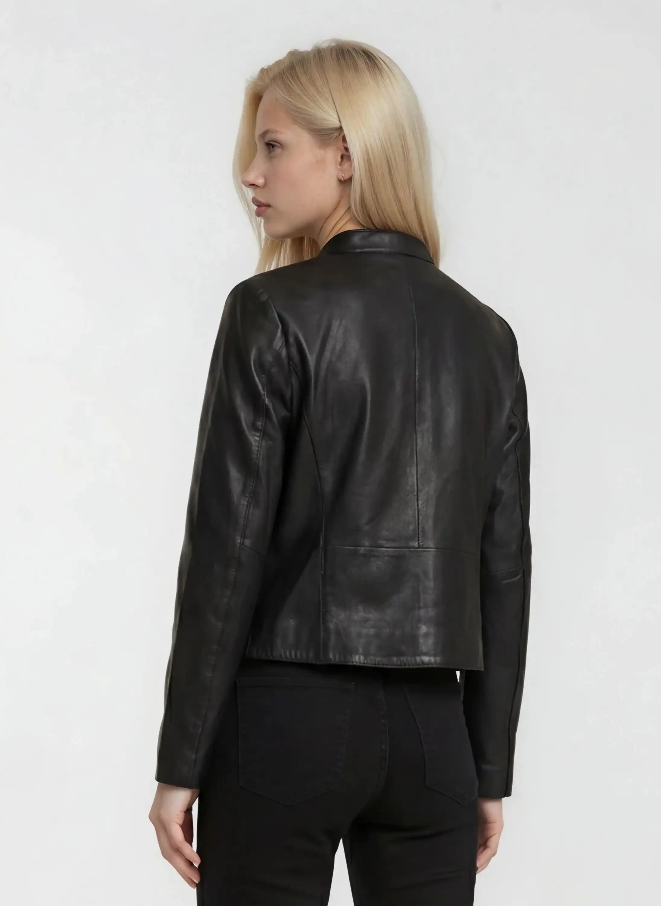 Eine Person mit langen blonden Haaren ist von hinten zu sehen. Sie trägt die trueprodigy Damen Lederjacke aus Schafsleder in Schwarz mit minimalistischem Design, gepaart mit einer schwarzen Lederhose vor einem schlichten hellen Hintergrund.