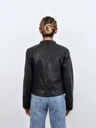 Eine Person mit blondem Haar in einem tiefen Dutt ist von hinten zu sehen. Sie trägt die trueprodigy Damen Lederjacke aus Schafsleder in Schwarz mit minimalistischem Design und hellblaue Jeans vor einem schlichten weißen Hintergrund.