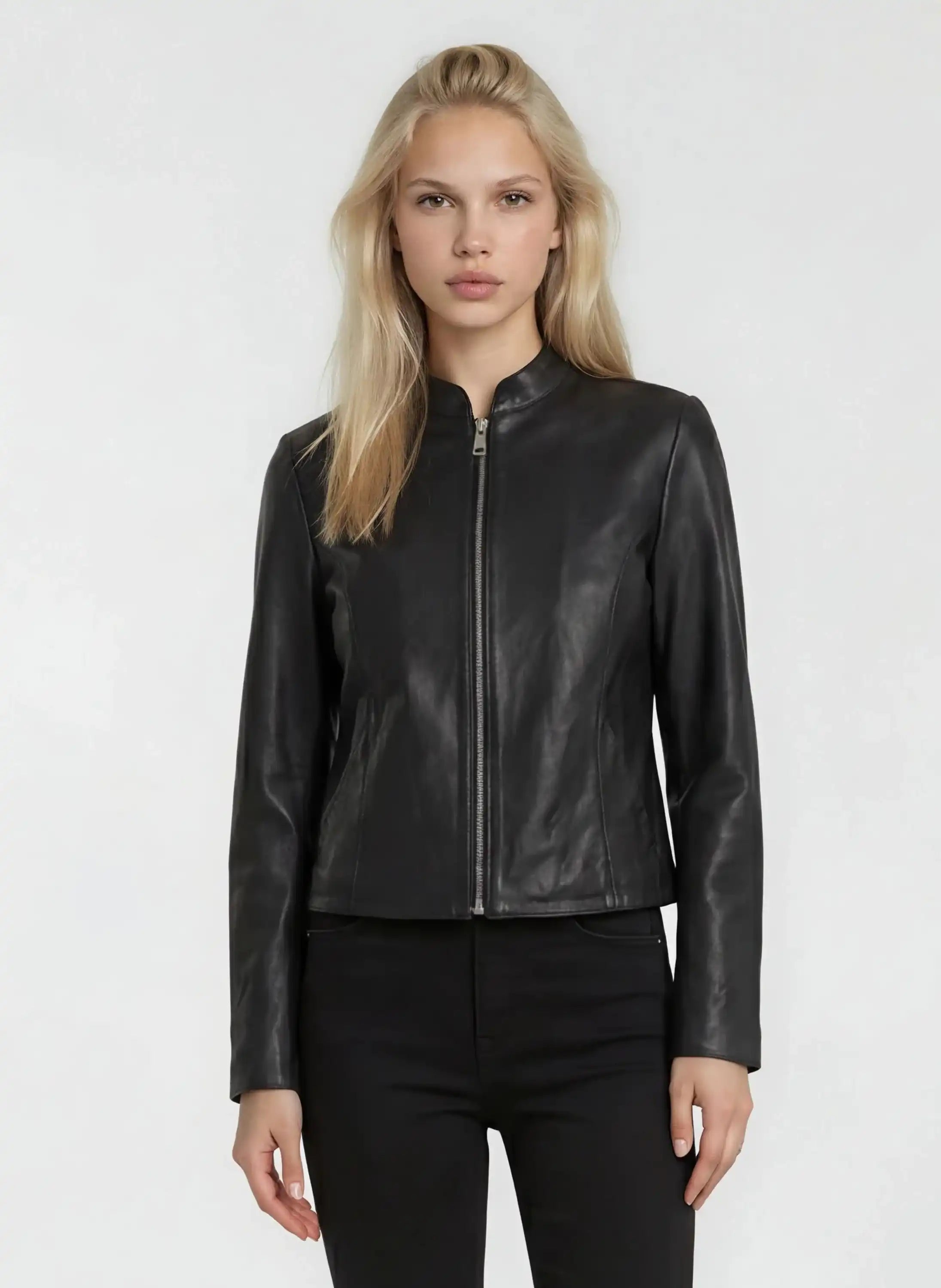 Eine Person mit langen blonden Haaren trägt die trueprodigy Damen Lederjacke aus Schafsleder in Schwarz mit minimalistischem Design und schwarzer Hose, stehend vor einem schlichten hellen Hintergrund.