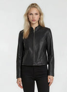 Eine Person mit langen blonden Haaren trägt die trueprodigy Damen Lederjacke aus Schafsleder in Schwarz mit minimalistischem Design und schwarzer Hose, stehend vor einem schlichten hellen Hintergrund.