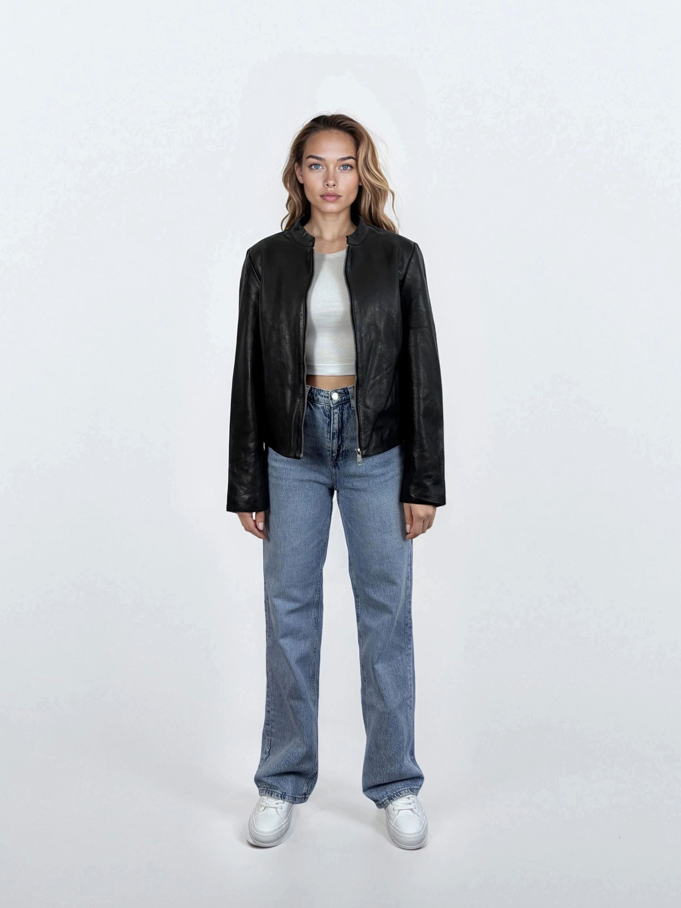 Eine Frau steht mit dem Gesicht nach vorne vor einem weißen Hintergrund und trägt eine trueprodigy Damen Lederjacke aus Schafsleder in Schwarz mit minimalistischem Design, gepaart mit einem weißen Crop-Top, blauen Jeans und weißen Sneakers. Ihre Arme sind an den Seiten verschränkt.
