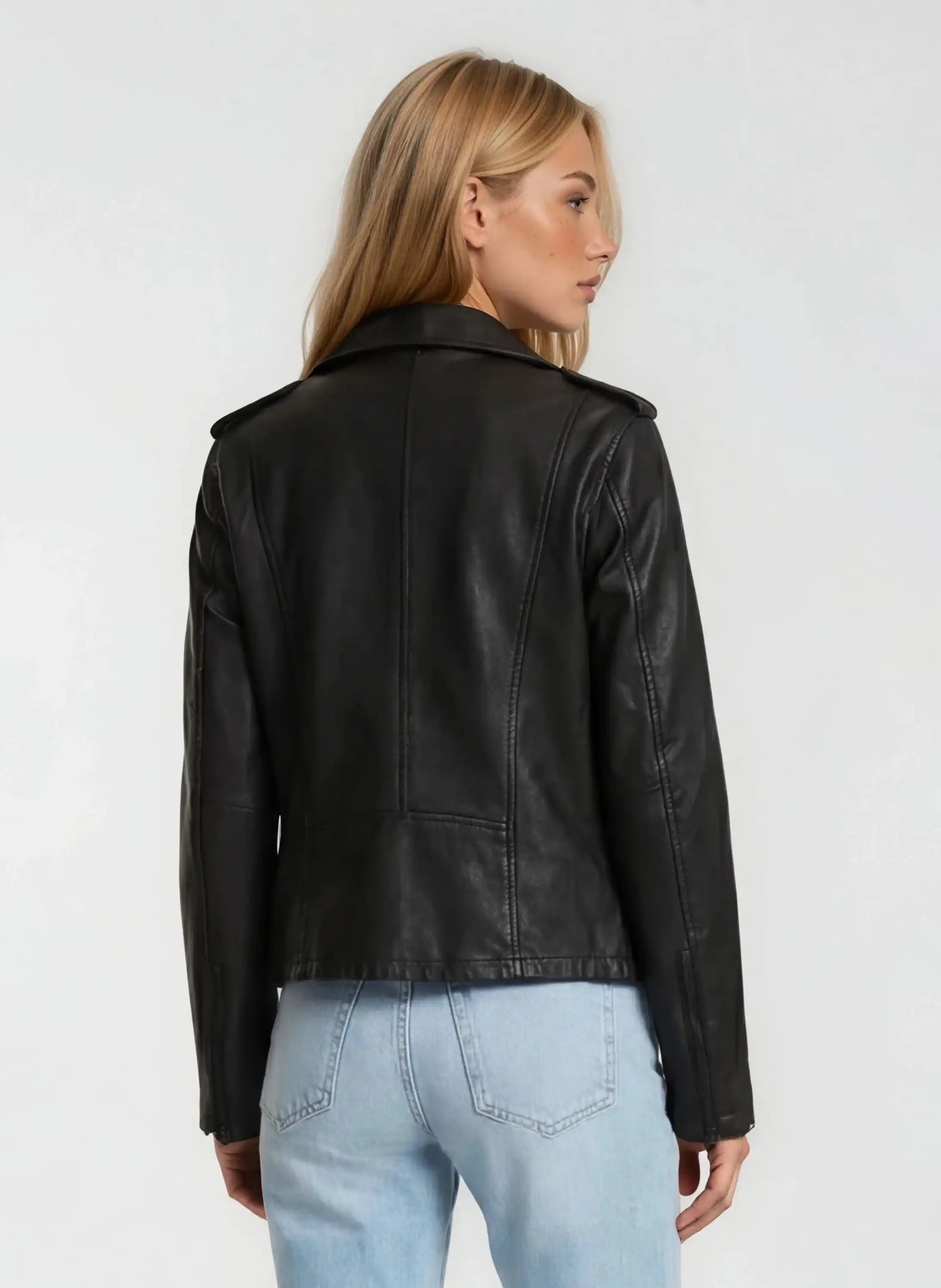 Man sieht eine Frau mit langen blonden Haaren von hinten, die die trueprodigy Damen Lederjacke aus Schafsleder in Schwarz im klassischen Biker-Stil und hellblaue Jeans vor einem schlichten hellen Hintergrund trägt.