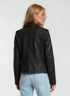 Man sieht eine Frau mit langen blonden Haaren von hinten, die die trueprodigy Damen Lederjacke aus Schafsleder in Schwarz im klassischen Biker-Stil und hellblaue Jeans vor einem schlichten hellen Hintergrund trägt.