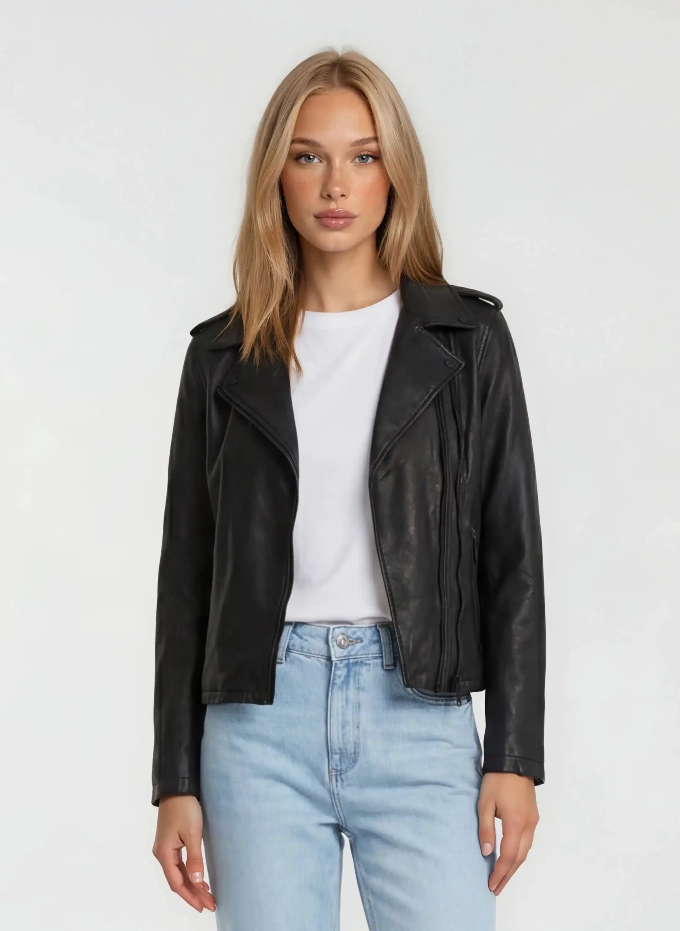 Eine Person mit glattem blondem Haar trägt eine trueprodigy Damen Lederjacke aus Schafsleder in Schwarz im klassischen Biker-Stil über einem weißen T-Shirt und hellblauen Jeans, die vor einem schlichten hellen Hintergrund stehen.