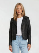 Eine Person mit glattem blondem Haar trägt eine trueprodigy Damen Lederjacke aus Schafsleder in Schwarz im klassischen Biker-Stil über einem weißen T-Shirt und hellblauen Jeans, die vor einem schlichten hellen Hintergrund stehen.