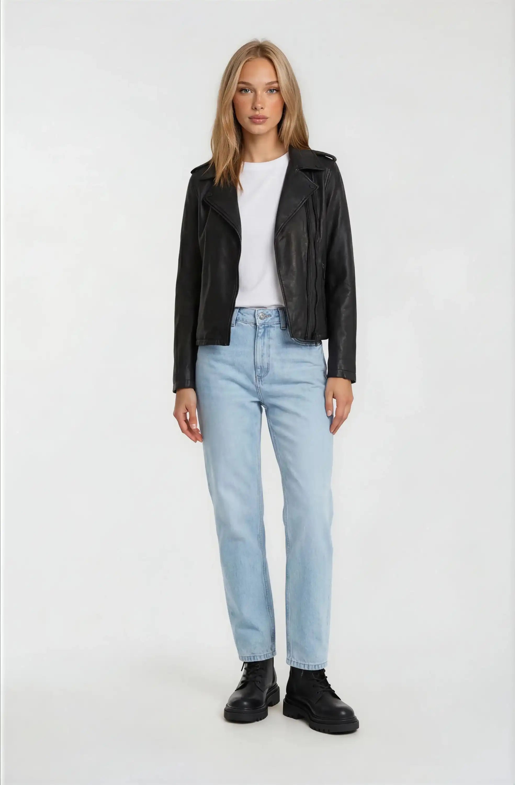 Eine Person steht vor einem weißen Hintergrund und trägt die trueprodigy Damen Lederjacke aus Schafsleder in Schwarz im klassischen Biker-Stil, ein weißes T-Shirt, hellblaue Jeans und schwarze Stiefel.