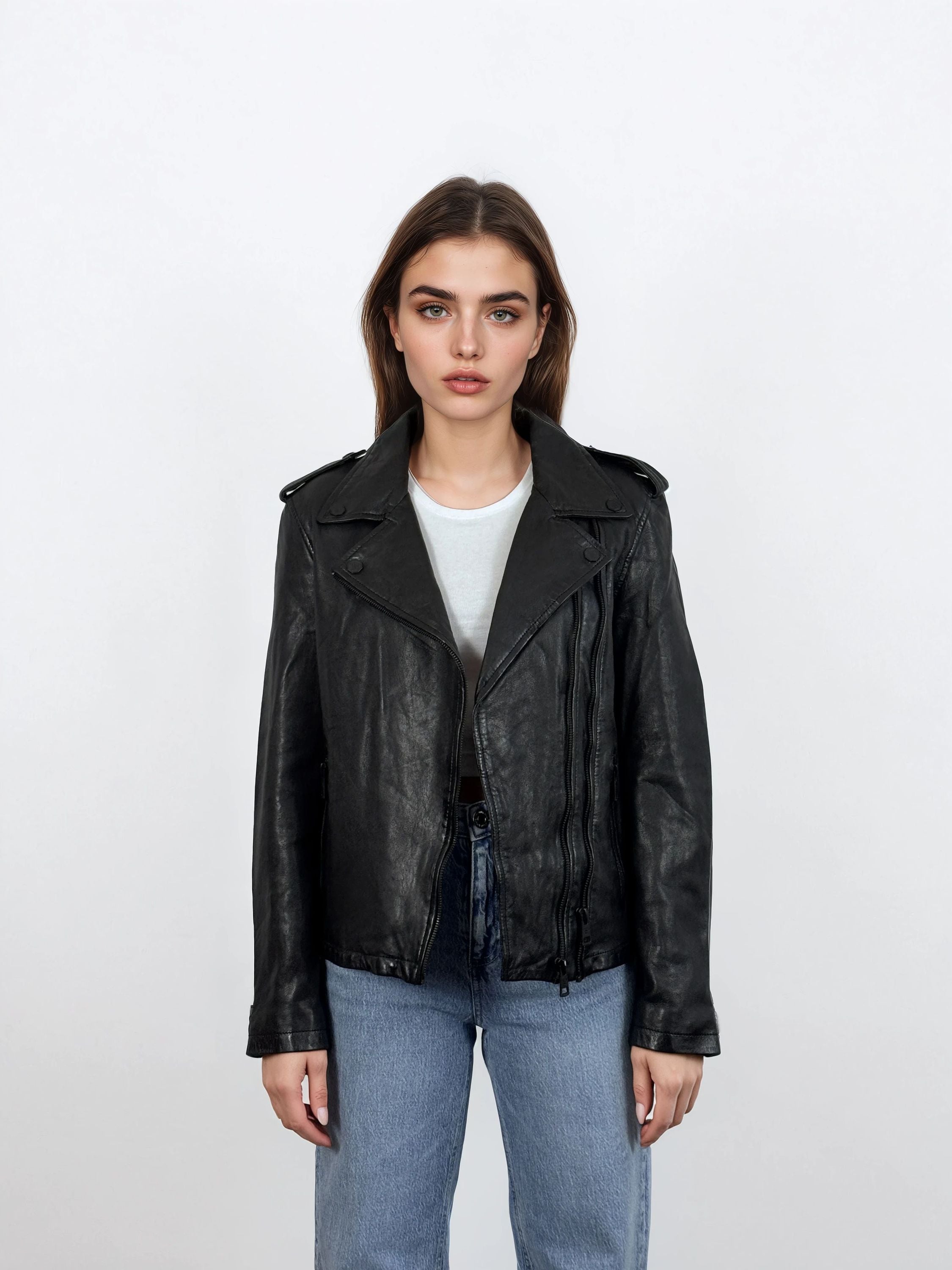 Eine Frau mit glattem braunem Haar trägt eine trueprodigy Damen Lederjacke aus Schafsleder in Schwarz im klassischen Biker-Stil über einem weißen T-Shirt und blauen Jeans und steht mit neutralem Gesichtsausdruck vor einem schlichten weißen Hintergrund.