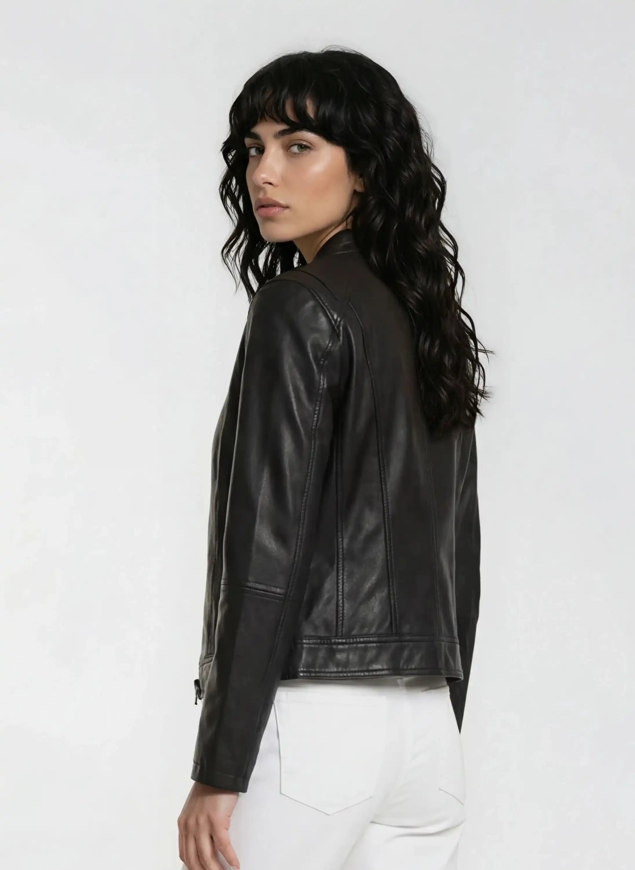 Eine Frau mit langen, gewellten dunklen Haaren trägt die trueprodigy Damen Lederjacke aus Schafsleder in Schwarz im modernen Café-Racer-Stil und eine weiße Hose, steht teilweise von der Kamera abgewandt vor einem hellen Hintergrund, der ihre weibliche Silhouette betont.