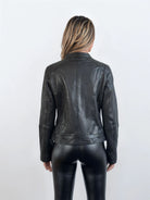 Eine Person mit langen, gewellten blonden Haaren wendet sich von der Kamera ab und zeigt eine feminine Silhouette in der trueprodigy Damen Lederjacke aus Schafsleder in Schwarz im modernen Café-Racer-Stil und passender Lederhose vor einem schlichten Hintergrund.