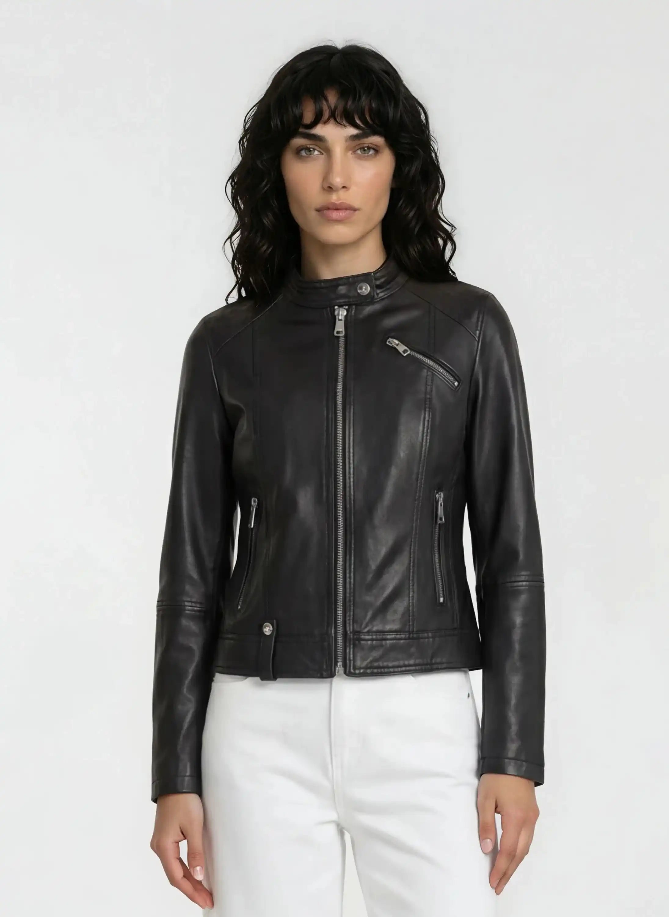 Eine Person mit langen lockigen dunklen Haaren trägt eine trueprodigy Damen Lederjacke aus Schafsleder in Schwarz im modernen Café-Racer-Stil mit Frontreißverschluss und Reißverschlusstaschen, gepaart mit einer weißen Hose, vor einem schlichten hellen Hintergrund.