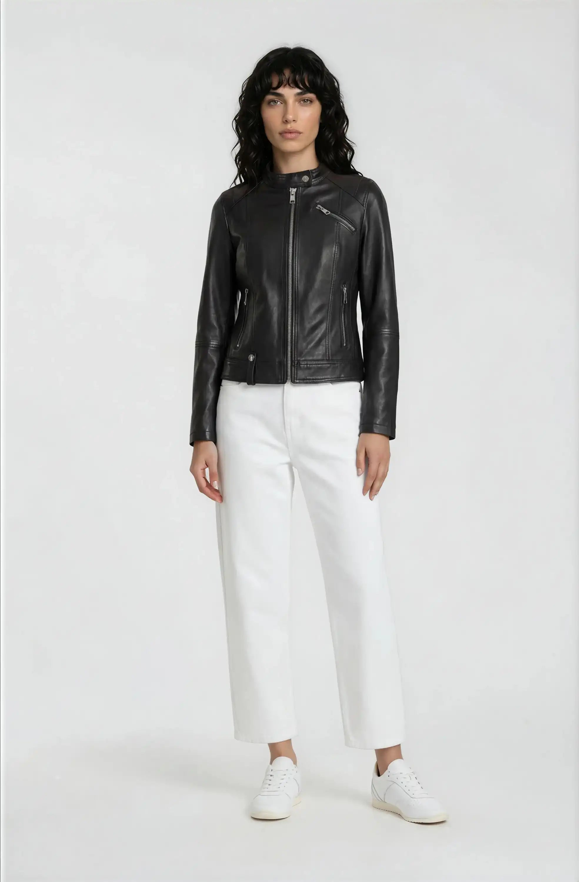 Eine Person steht vor einem schlichten Hintergrund und trägt die trueprodigy Damen Lederjacke aus Schafsleder in Schwarz im modernen Café-Racer-Stil, gepaart mit einer weißen, weit geschnittenen Hose und Sneakers. Sie hat dunkles, gewelltes Haar und eine moderne, feminine Silhouette.