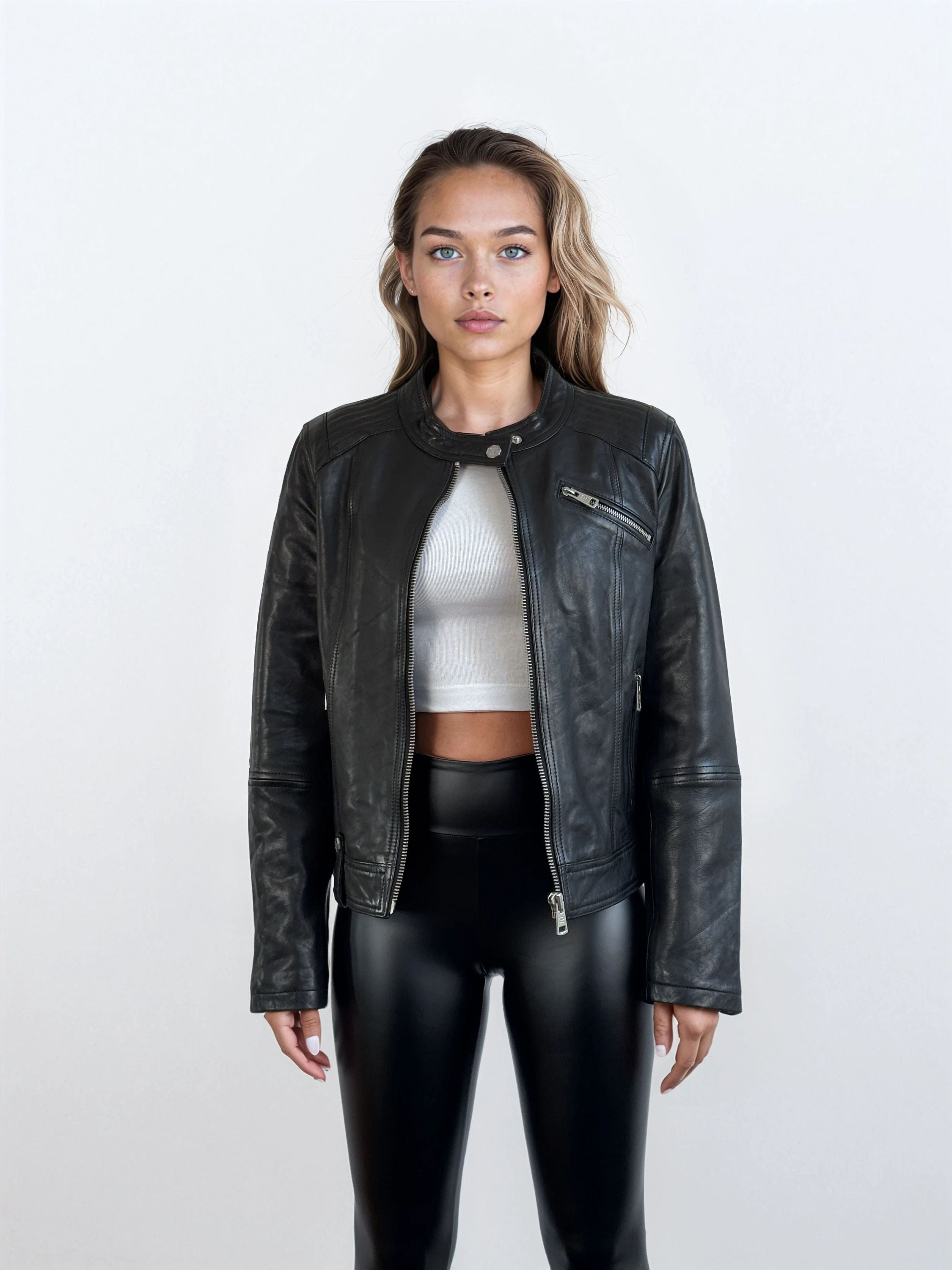 Eine Frau trägt die schwarze Damen Lederjacke aus Schafsleder im modernen Café-Racer-Stil von trueprodigy über einem weißen Crop-Top und glänzenden schwarzen Hosen vor einem schlichten hellen Hintergrund.