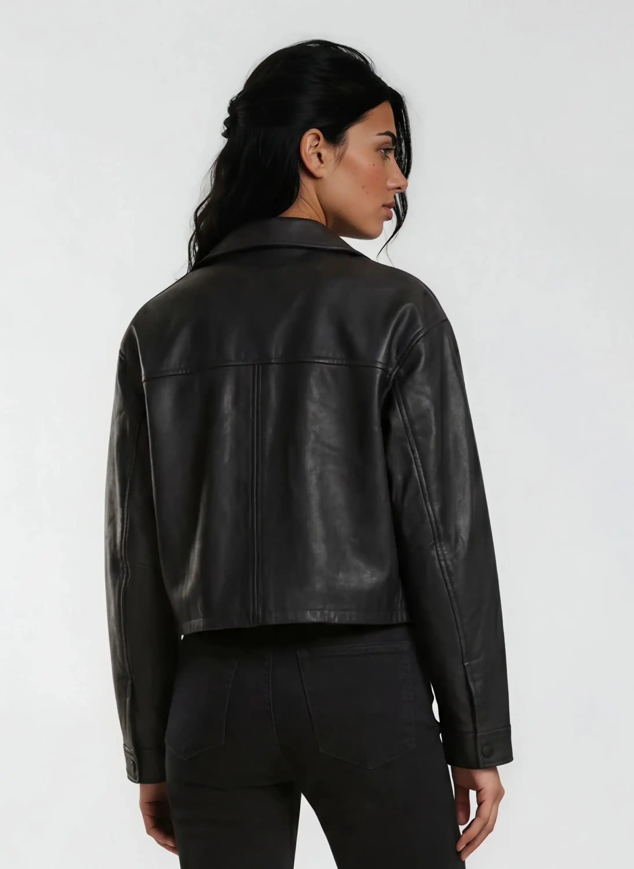 Eine Person mit langen dunklen Haaren trägt die trueprodigy Damen Lederjacke aus Schafsleder in Schwarz im Oversized Cropped-Style und schwarze Jeans und steht mit dem Rücken zur Kamera vor einem schlichten hellen Hintergrund.