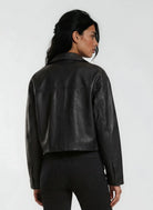 Eine Person mit langen dunklen Haaren trägt die trueprodigy Damen Lederjacke aus Schafsleder in Schwarz im Oversized Cropped-Style und schwarze Jeans und steht mit dem Rücken zur Kamera vor einem schlichten hellen Hintergrund.