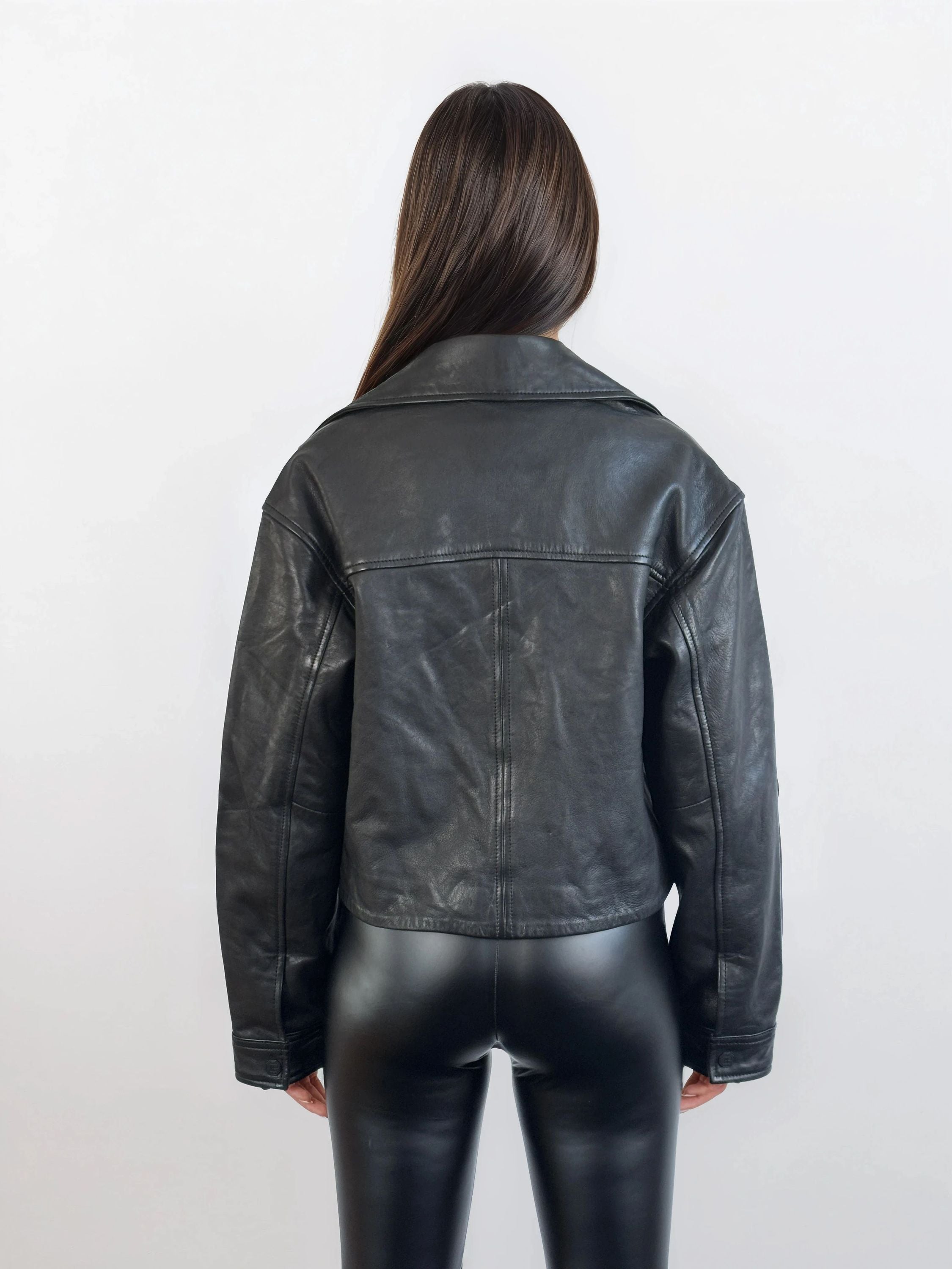 Eine Frau mit langen braunen Haaren steht mit der trueprodigy Damen Lederjacke aus Schafsleder in Schwarz im Oversized Cropped-Style und einer glänzenden schwarzen Hose vor einem schlichten hellen Hintergrund.