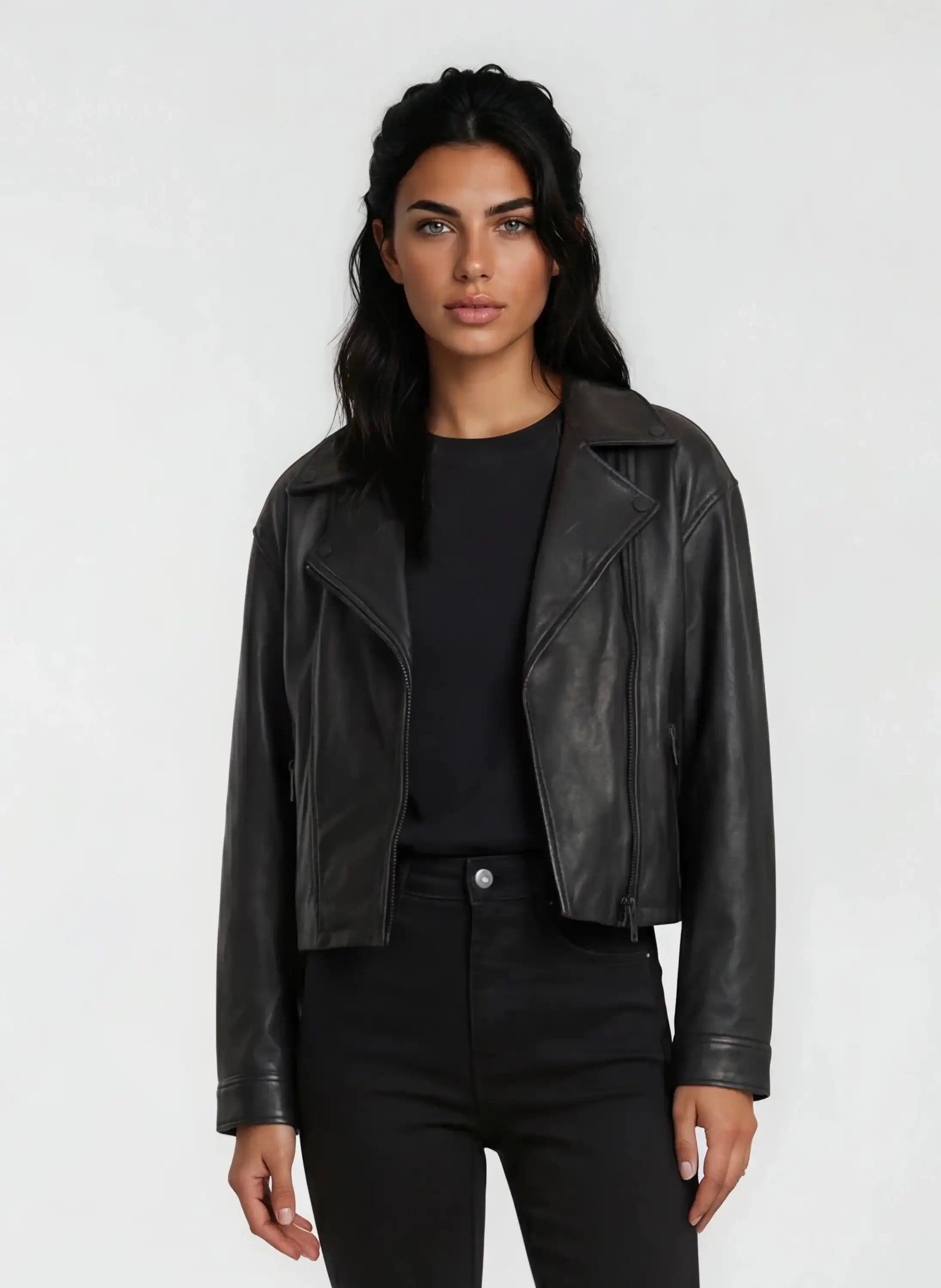 Eine Frau mit langen dunklen Haaren trägt die trueprodigy Damen Lederjacke aus Schafsleder in Schwarz im Oversized Cropped-Style über einem schwarzen Hemd und einer Hose und steht mit dem Gesicht nach vorne vor einem schlichten hellen Hintergrund.