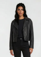 Eine Frau mit langen dunklen Haaren trägt die trueprodigy Damen Lederjacke aus Schafsleder in Schwarz im Oversized Cropped-Style über einem schwarzen Hemd und einer Hose und steht mit dem Gesicht nach vorne vor einem schlichten hellen Hintergrund.