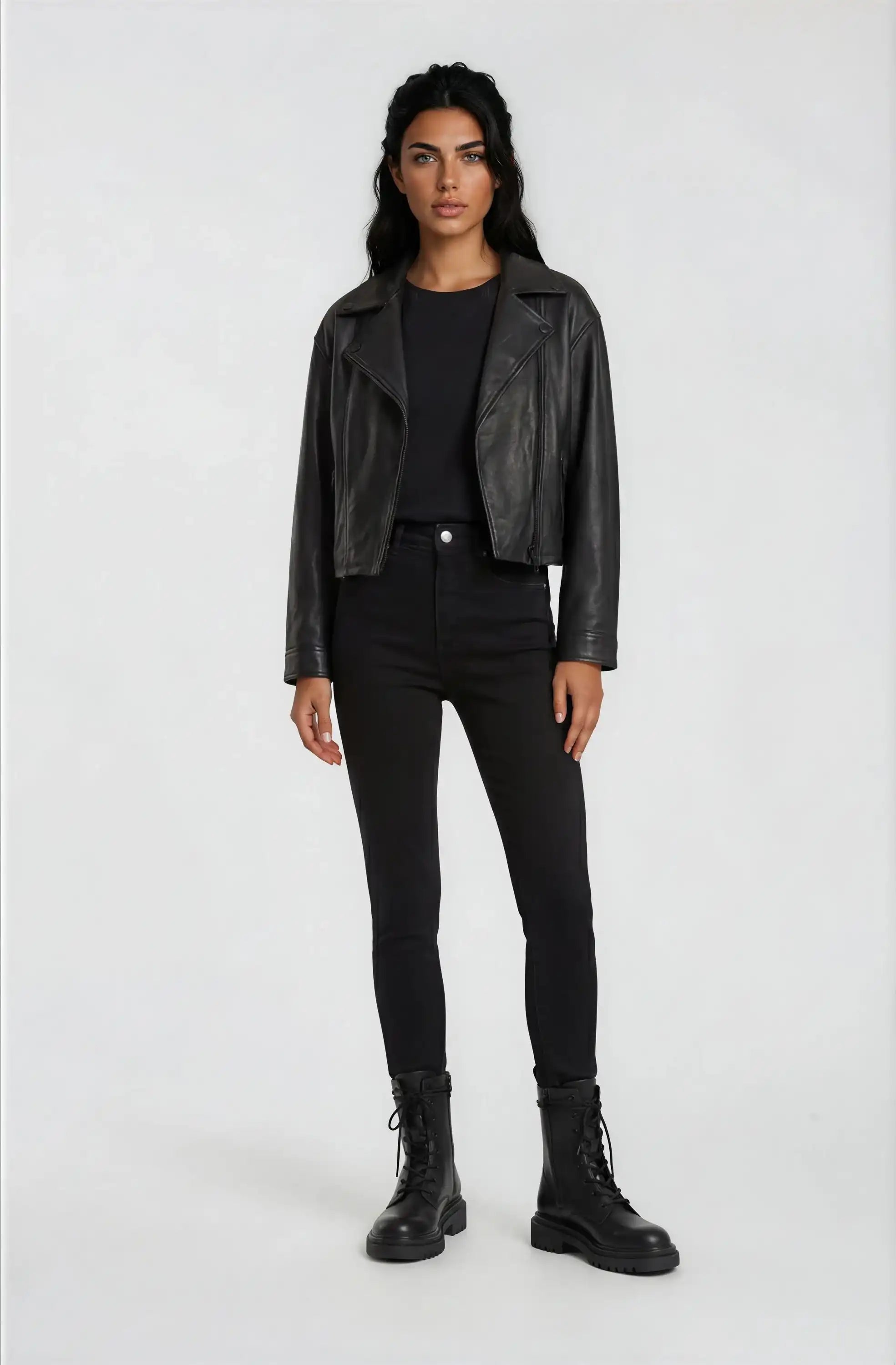 Eine Person steht mit dem Gesicht nach vorne vor einem unifarbenen Hintergrund und trägt eine trueprodigy Damen Lederjacke aus Schafsleder in Schwarz im Oversized Cropped-Style, ein schwarzes T-Shirt, High-Waisted Skinny Jeans und schwarze Schnürstiefel.