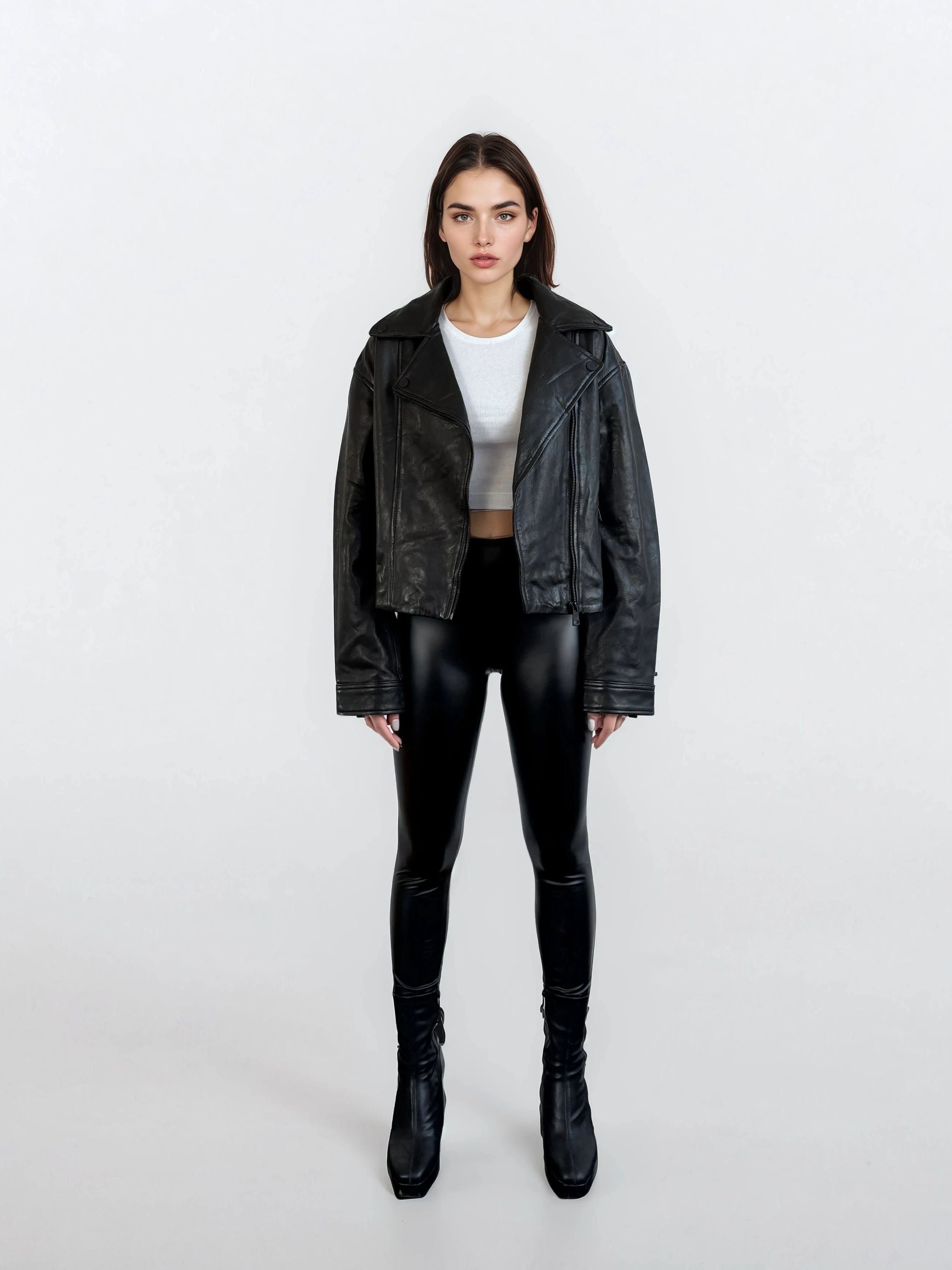 Eine Frau steht vor einem weißen Hintergrund und trägt die trueprodigy Damen Lederjacke aus Schafsleder in Schwarz im Oversized Cropped-Style, gepaart mit einem weißen Cropped-Top, schwarzen, glänzenden Leggings und Stiefeln mit Absatz und blickt neutral in die Kamera.