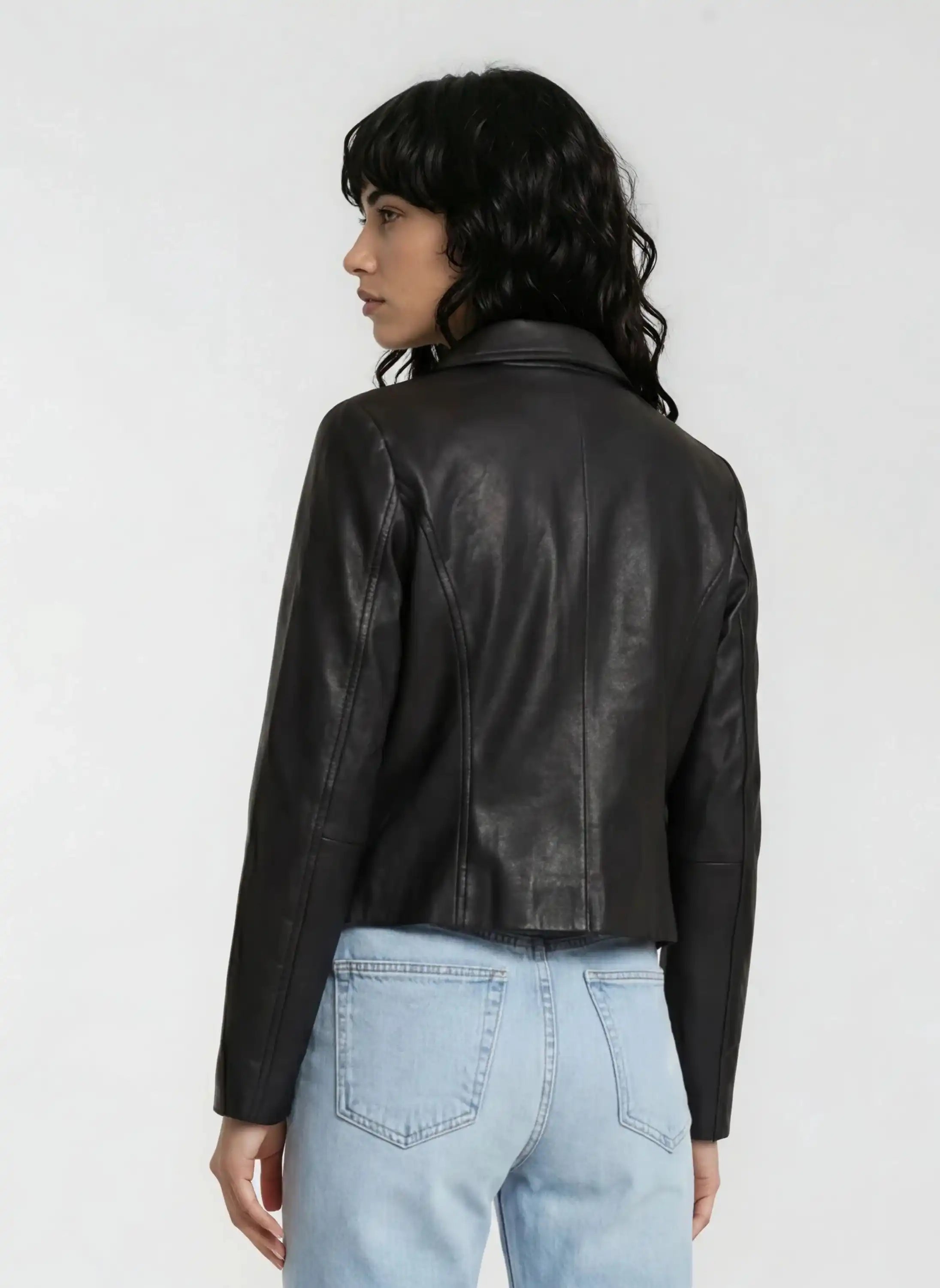 Eine Person mit langem, dunklem, welligem Haar trägt vor hellem Hintergrund eine schwarze trueprodigy Damen Lederjacke aus Schafsleder mit minimalistischem Design und hellblauen Jeans im Blouson-Stil.